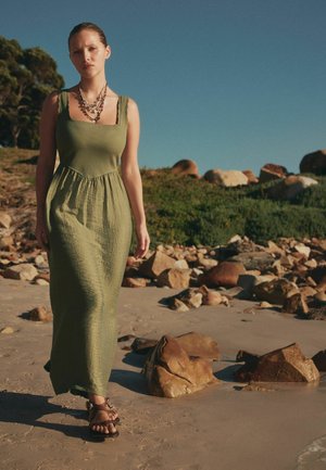 Kvinde går barfodet på en sandstrand iført en olivengrøn ærmeløs maxikjole og lagdelte halskæder, med klipper og grønt i baggrunden.