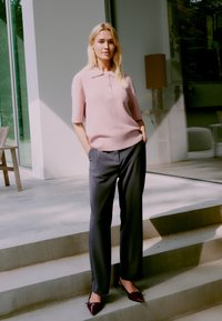 Pull court rose clair à manches courtes avec trois boutons, associé à un pantalon gris foncé ajusté et des talons aiguilles bordeaux, se tenant sur des marches en pierre.