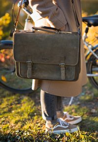 Bolsa de mensajero de cuero marrón con dos correas frontales, un asa y una correa de hombro, apoyada contra el costado de una persona al aire libre.