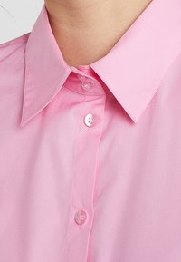Camicia di cotone rosa con colletto a punta, dotata di abbottonatura frontale e superficie liscia. I bottoni sono in plastica trasparente, offrendo dettagli sottili.