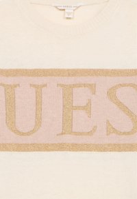 Krémový a světle růžový svetr s výrazným zlatým logem "GUESS" napříč hrudníkem. Měkká textura se žebrovaným výstřihem.