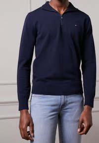 Pull-over bleu marine avec col zippé, poignets et ourlet côtelés. Texture lisse, coupe ajustée, avec un petit logo sur la poitrine.