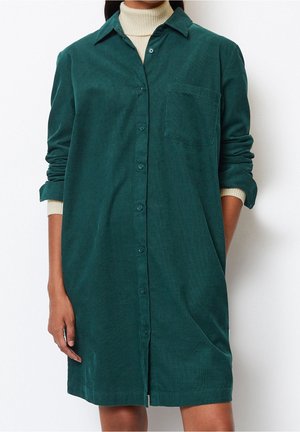 Woman wearing a dark green corduroy shirt dress with long sleeves rolled up, buttoned front, and a beige turtleneck underneath.