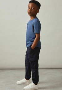 T-shirt en coton bleu, à manches courtes, associé à un pantalon cargo sombre en marine avec des poches, et des baskets blanches, se tenant devant un arrière-plan neutre.