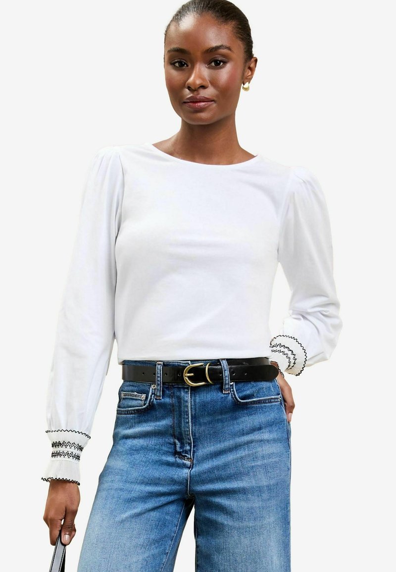 Maglietta bianca a maniche lunghe con spalle arricciate, rifinita con bordini neri ricamati sui polsini, abbinata a jeans in denim a vita alta e una cintura nera.