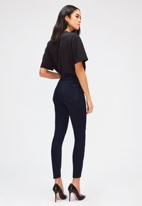 T-shirt noir oversize associé à un jean skinny bleu foncé taille haute. Le modèle porte des chaussures à talons hauts noires. Arrière-plan épuré et minimaliste.