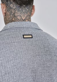 Camicia grigia a spina di pesce con colletto, caratterizzata da un'etichetta nera decorativa sul retro e da un motivo tessuto texturizzato.