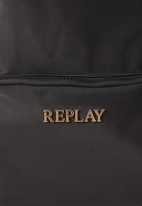Sac en tissu noir avec le logo "REPLAY" en lettres dorées et une fermeture éclair horizontale au-dessus du logo.