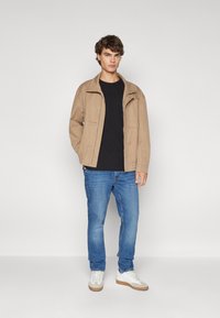 Chaqueta beige hecha de material suave, con cremallera frontal, dos bolsillos laterales, llevada sobre una camisa negra, con jeans azules y zapatillas blancas.
