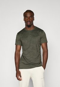 Olive green short-sleeve t-shirt with a camouflage pattern, crew neckline, made of smooth fabric, fitted design, worn with light-colored pants.