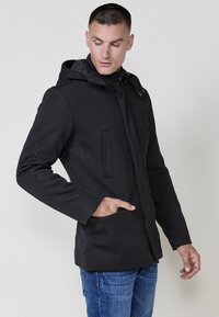 Giacca nera con cappuccio e finitura testurizzata, dotata di chiusura con zip e due tasche anteriori. Indossata sopra jeans blu, mette in risalto una vestibilità slim.