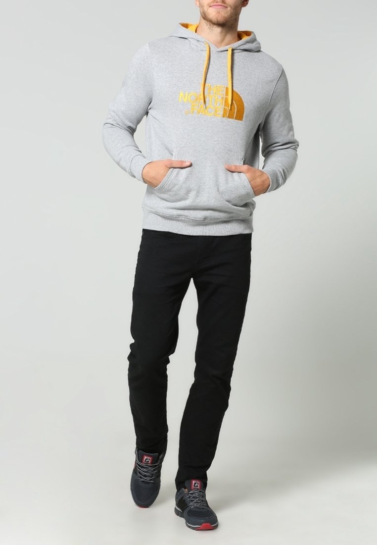 Homme portant un sweat à capuche gris avec un logo jaune et des cordons, un pantalon noir et des baskets foncées, debout les mains dans la poche du sweat.