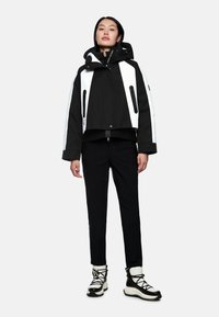 Gekürzte schwarz-weiße Jacke mit Kapuze, die mit Reißverschlusstaschen und kontrastierenden weißen Einsätzen ausgestattet ist. Kombiniert mit schwarzen Hosen und klobigen Stiefeln.