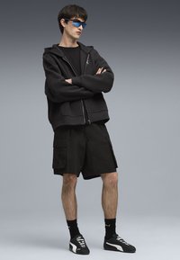 Felpa nera con zip e vestibilità rilassata, abbinata a pantaloni cargo neri, completata da occhiali da sole blu e scarpe sportive nere.