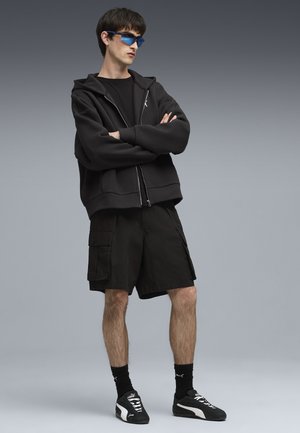Sort zip-up hættejakke i afslappet pasform, parret med sorte cargo shorts, suppleret med blå solbriller og sorte sportssko.