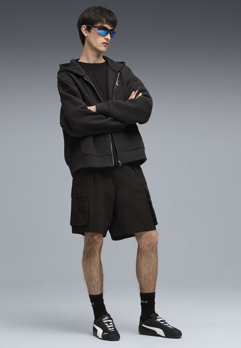 Sudadera negra con cremallera y un corte relajado, combinada con pantalones cortos cargo negros, complementada con gafas de sol azules y zapatillas deportivas negras.