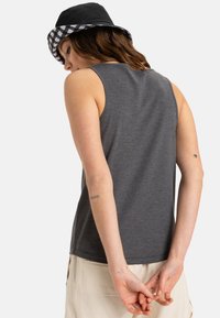Top grigio senza maniche con una texture liscia e scollatura rotonda, indossato con un cappello a secchiello a quadri bianchi e neri. Mani unite dietro la schiena.