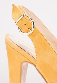 Žlutá semišová lodička na vysokém podpatku s špičatou špičkou a designem s otevřenou patou. Má stříbrnou sponu a několik nastavovacích otvorů.