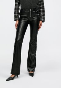 Μαύροι παντελόνι από ψεύτικο δέρμα με φερμουάρ σε τσέπες μπροστά, λεία υφή και εφαρμοστή γραμμή. Φορεμένο με πουκάμισο καρό και μαύρες ψηλοτάκουνες γόβες.