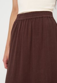 Personne portant un haut crème sans manches et une jupe marron ample taille haute avec une ceinture élastique, visible de la taille au milieu des cuisses.