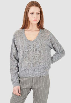 Donna con capelli rossi lisci che indossa un maglione a V grigio chiaro con motivo e pantaloni grigi a righe verticali, in piedi con una mano sui fianchi.