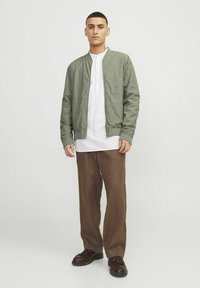 Grüne Bomberjacke über einem weißen Henley-Shirt, kombiniert mit braunen lässigen Hosen und dunklen Schuhen. Glatter Stoff, minimalistisches Design.