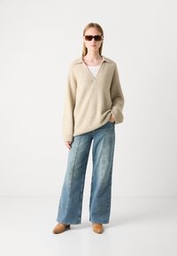 Beige, fluffy v-hals sweater med løs pasform, parret med brede, lyseblå denim jeans og brune ankelstøvler. Brune solbriller fremhæver looket.