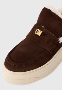 Pantofii slip-on din suede maro închis, cu talpă din cauciuc crem, căptușiți cu fleece și logo-ul auriu "SM" pe brățara de deasupra.