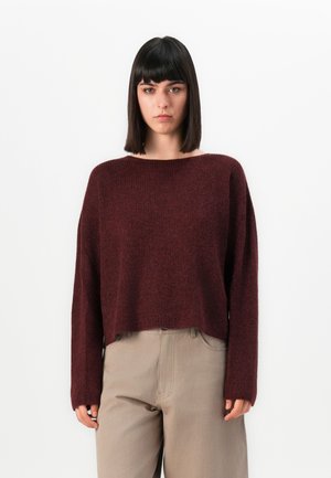 Kvinne med rett svart hår iført en løs maroon genser og beige bukser, står mot en enkel hvit bakgrunn.