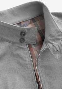 Chaqueta gris con un alto cuello, que presenta un cierre frontal, dos botones negros y un forro de cuadros en tonos marrones y rojos.