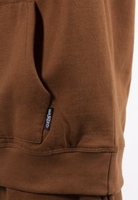 Felpa marrone in pile con tasca a marsupio, polsini a coste e un piccolo etichetta nera con la scritta "WASTED" sul lato sinistro. Texture morbida.