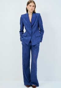 Costume texturé bleu avec un motif floral, comprenant un blazer ajusté avec des revers en pointe et un pantalon à jambes larges. Boutons et poches inclus.