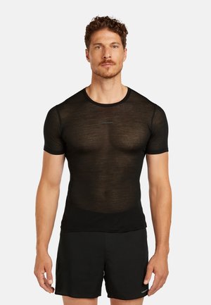 Mężczyzna w dopasowanej czarnej siateczkowej koszulce z krótkim rękawem i czarnych szortach, stojący na jasnym, jednolitym tle, zwrócony przodem.