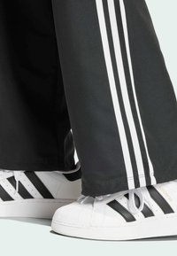 Svarta byxor med vita tre-strimmiga accenter, tillsammans med vita sneakers med svarta ränder och texturerad tåkuppel.