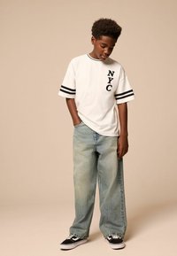 Weißes T-Shirt mit kurzen Ärmeln und schwarzen Akzenten sowie dem Text "NYC", kombiniert mit hellblauen weiten Jeans und schwarzen Sneakers. Flüssige Stofftextur.