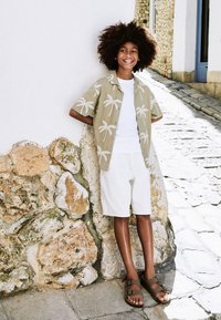 Camicia verde chiaro a maniche corte con motivo di palme bianche, t-shirt bianca, pantaloncini beige e sandali marroni, in piedi vicino a un muro di pietra.