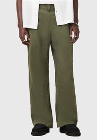 Pantalons à jambes larges vert olive avec une texture lisse, comportant des détails de panneaux et une taille haute. Portés avec des chaussures noires à enfiler.