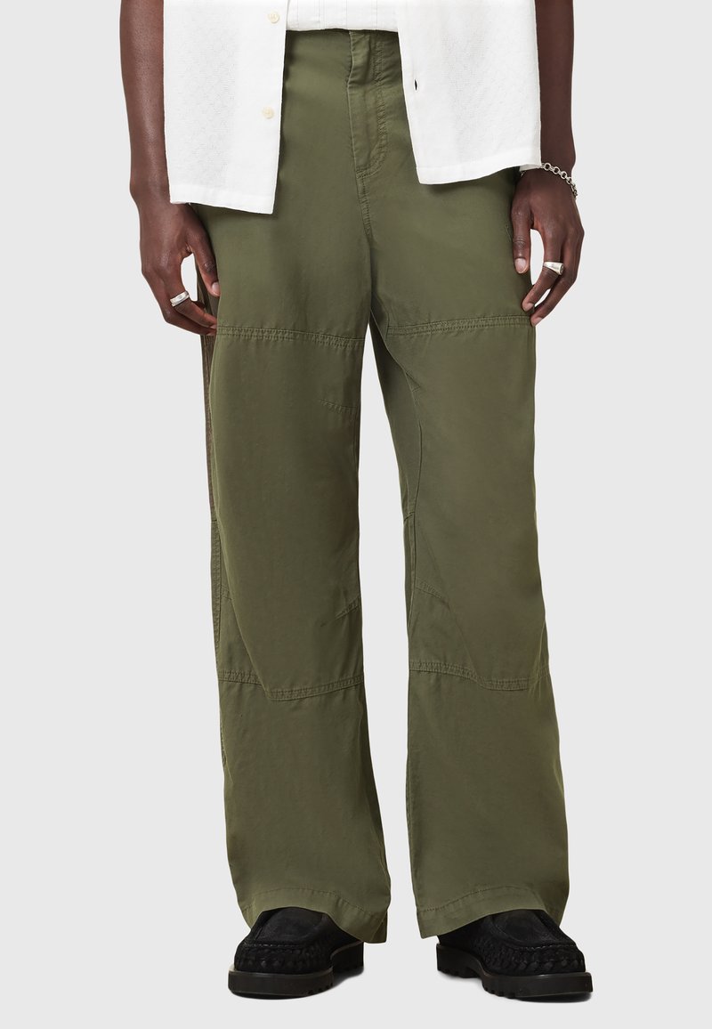 Pantalons à jambes larges vert olive avec une texture lisse, comportant des détails de panneaux et une taille haute. Portés avec des chaussures noires à enfiler.