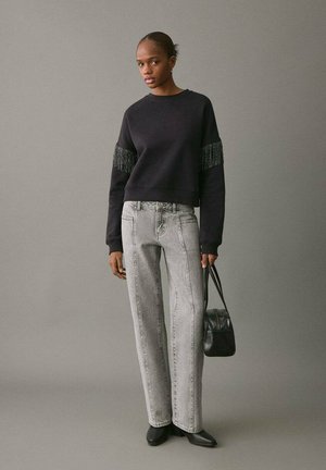 Schwarzer Pullover mit strukturierten Fransensäumen, kombiniert mit hellgrauen geraden Jeans. Das Modell hält eine schwarze Tasche und trägt spitze schwarze Schuhe.