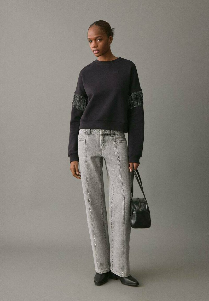 Svart sweatshirt med texturerade fransade ärmar, parat med ljusgrå raka jeans. Modellen håller en svart väska och bär spetsiga svarta skor.