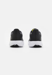 Zapatillas deportivas negras con parte superior de malla texturizada y suela blanca. La vista trasera muestra un acento amarillo en el talón y un diseño ligero.