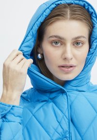 Modrá izolačná puffáčka s prešívaným vzorom, s kapucňou a zapínaním na cvočky. Tkanina vyzerá hladko s lesklou textúrou.
