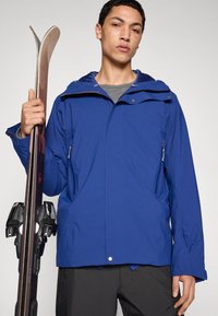 Blå skidjacka med huva, utrustad med dragkedjeförsedda fickor, slät yta och kombinerad med skidor hållna i höger hand.