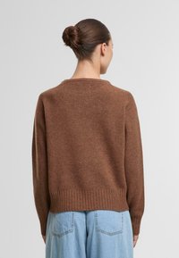 Brauner Strickpullover mit geripptem Saum, lockerer Passform und Rundhalsausschnitt. Über hellblauen weit geschnittenen Hosen getragen, die einen texturierten Stoff zeigen.