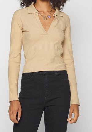 Beige ribbet langærmet top med krave og V-udskæring, parret med sorte jeans. Guld kædehalssmykke synligt på halsen.