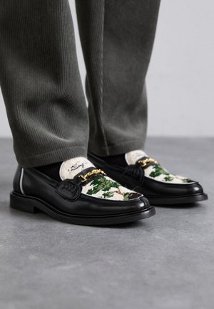 Mocassins de couro preto com um superiores têxtil decorativo verde e branco. Apresenta um detalhe de corrente dourada e sola preta texturizada.