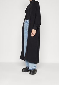 Cardigan lungo nero in maglia con vestibilità comoda, abbinato a jeans azzurri con orlo risvoltato, indossato con calzature nere chunky.