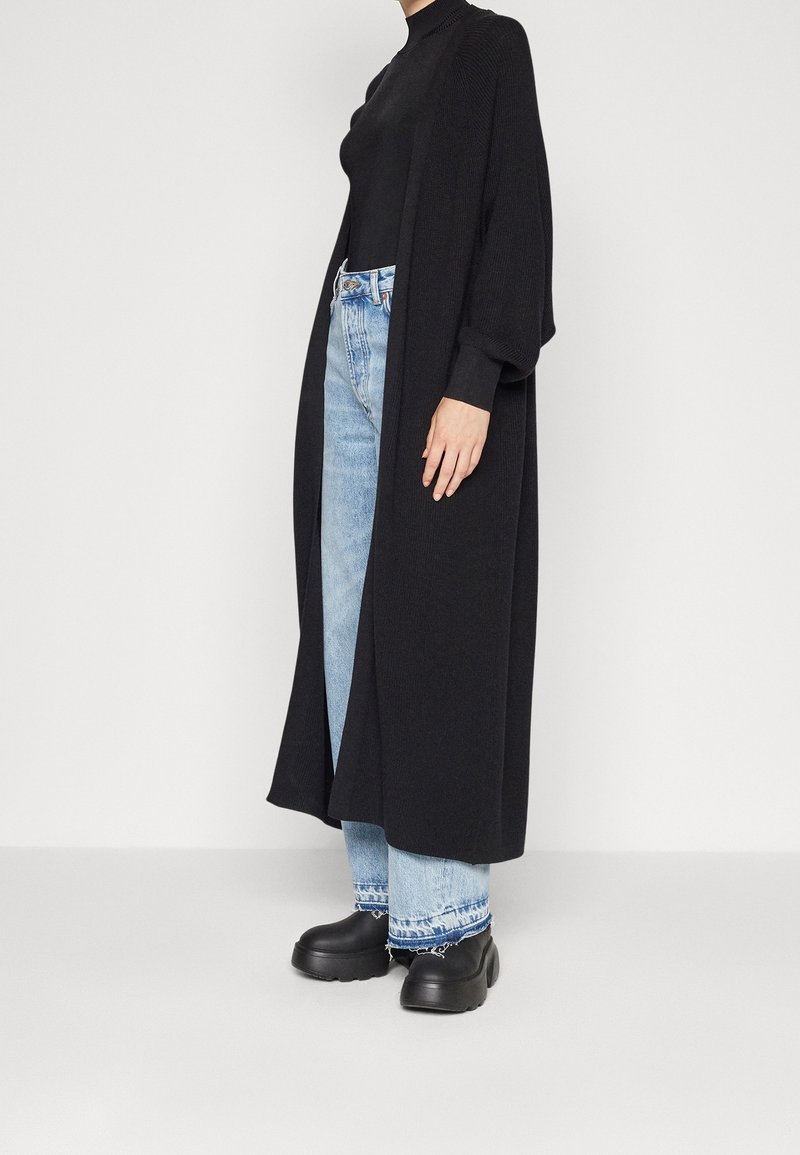 Cardigan lungo nero in maglia con vestibilità comoda, abbinato a jeans azzurri con orlo risvoltato, indossato con calzature nere chunky.