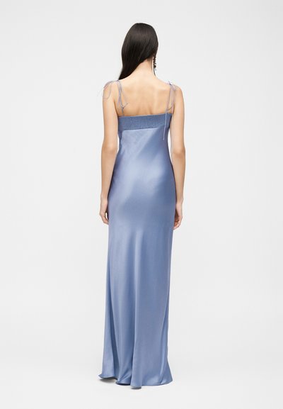 Γυναίκα με μακριά σκούρα μαλλιά, φορώντας ένα μακρύ σατέν φόρεμα ανοιχτού μπλε χρώματος με λεπτές τιράντες, φαίνεται από πίσω με φόντο ένα μονόχρωμο υπόβαθρο.