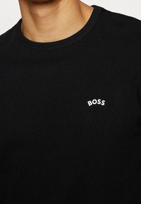 Homme portant un t-shirt noir à col rond avec le mot "BOSS" imprimé en blanc sur le côté gauche de la poitrine.